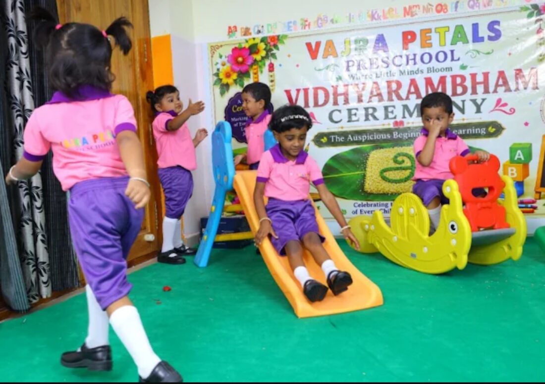 Children in pink and purple uniforms playing on an indoor slide beneath the Vajra Petals Vidhyarambham ceremony banner